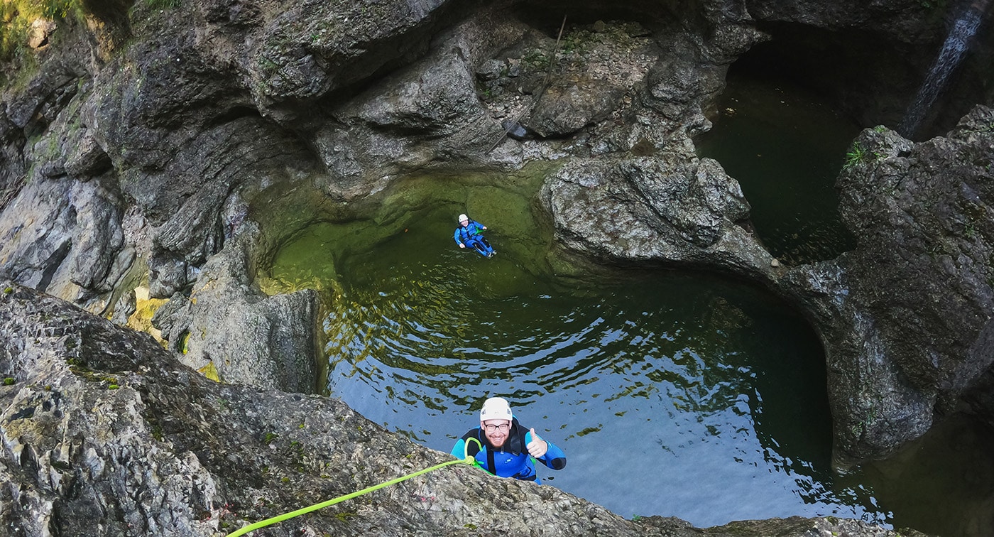 Zwei Personen bei einem Outdoor-Abenteuer mit mehreren Aktivitäten in der Almbachklamm. Eine Person in einem natürlichen Wasserbecken, während die andere sicher an einem Seil gesichert nach oben klettert. Probier Canyoning in Salzburg, während du in Österreich bist.
