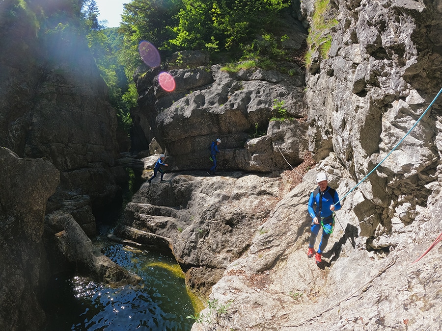 Gruppe von Erwachsenen beim Canyoning, die an einem Seil gesichert an Felsen entlang gehen, in der Almbachklamm (Wiestal) Salzburg, an einem sonnigen Tag.